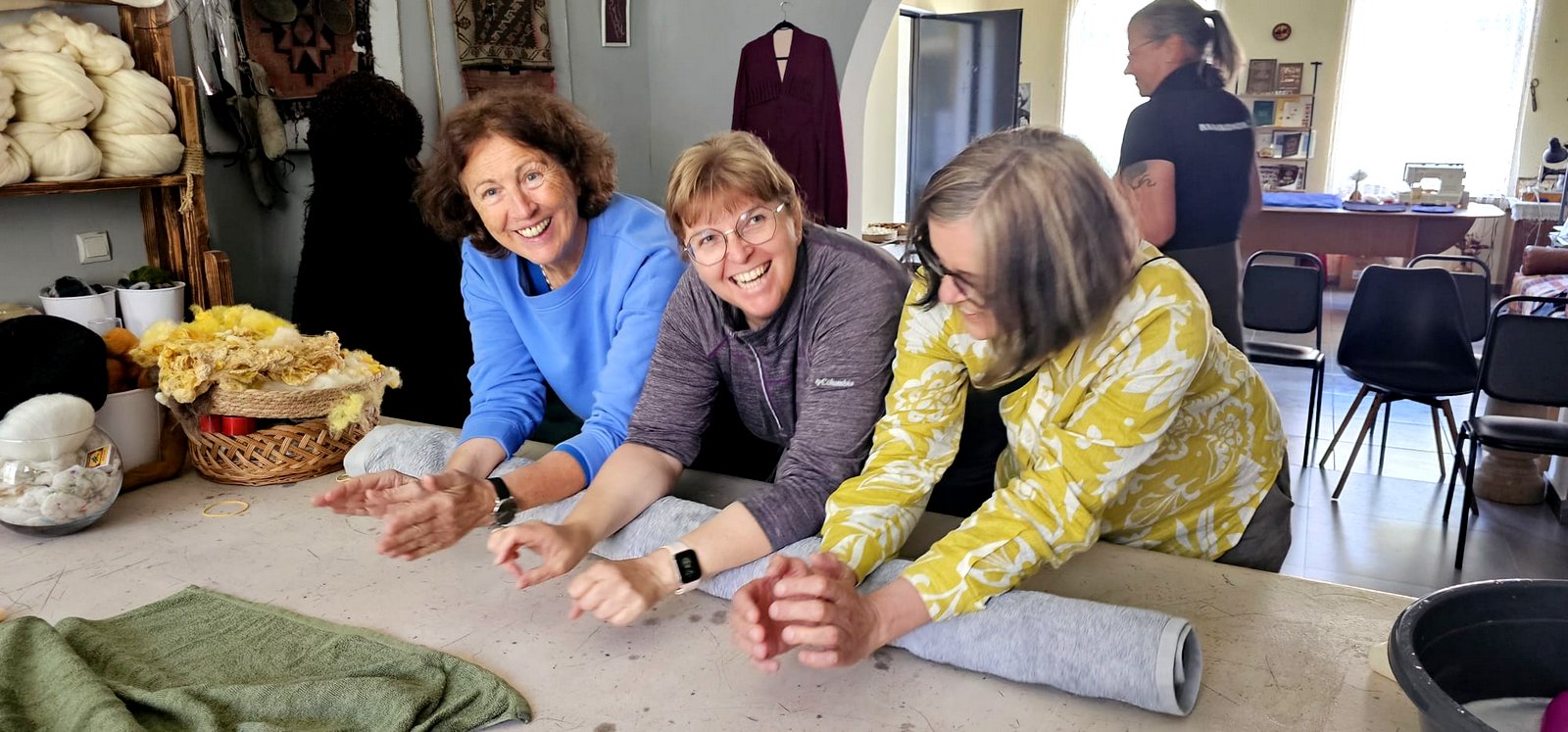 Drei lachende Frauen beim gemeinsamen Filzen an einem Arbeitstisch in einer georgischen Filzwerkstatt, Wollmaterialien im Hintergrund