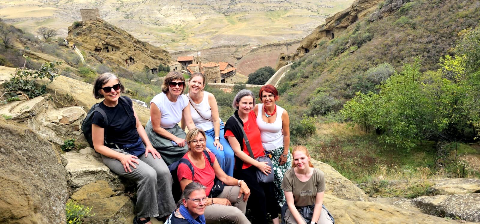 Reisegruppe sitzt auf Felsen oberhalb des Höhlenklosters David Gareji in der georgischen Steppe, im Hintergrund die Klosterbauten im Tal