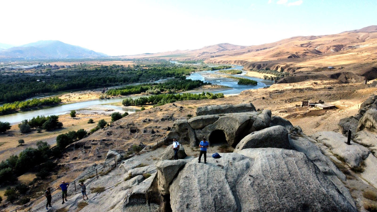 Blick von der antiken Höhlenstadt Uplistsikhe über den Fluss Mtkvari in der zentralgeorgischen Steppe – in den Fels gehauene Kammern im Vordergrund, Besucher auf dem Felsplateau