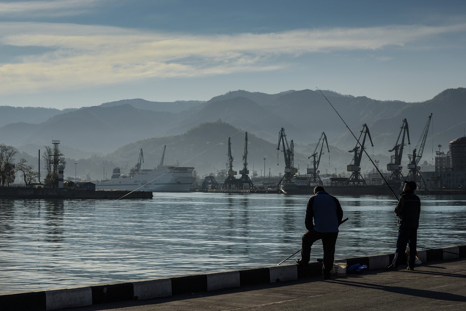 Zwei Männer angeln im Gegenlicht am Hafenkai von Batumi vor Hafenkränen Schiffen und Bergen im Morgendunst am Schwarzen Meer