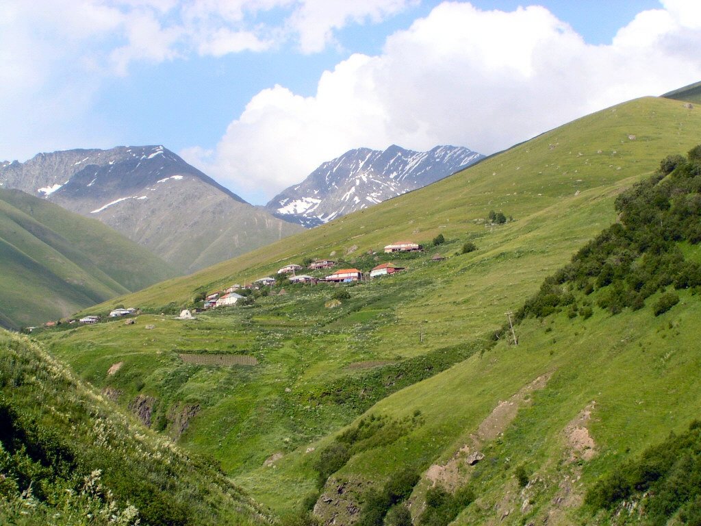 Das kleine Bergdorf Dshuta im Sno-Tal mit grünen Hängen und schneebedeckten Gipfeln des Chaukhi-Massivs im Hintergrund