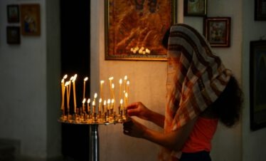 Eine Frau mit Kopftuch zündet Kerzen vor einer Ikone in einer georgisch-orthodoxen Kirche an warmem Kerzenlicht