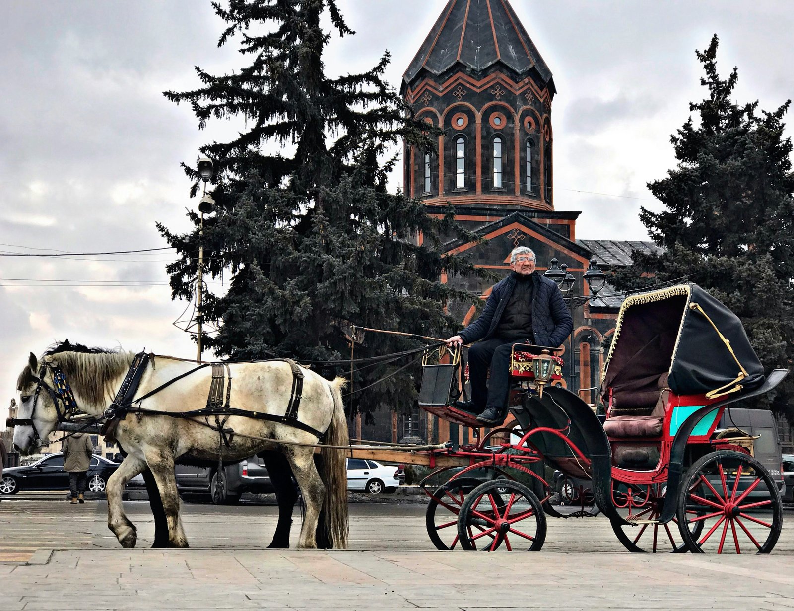 Ein älterer armenischer Mann auf einer roten Pferdekutsche mit einem weißen Pferd vor einer Kirche aus rot-schwarzem Vulkanstein in Gjumri