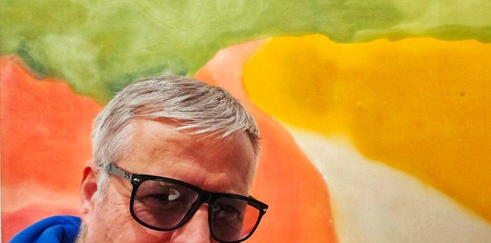 Hans Heiner Buhr, Künstler und Inhaber von Kaukasus-Reisen, posiert vor einem großformatigen, abstrakten Kunstwerk in kräftigen Rot-, Gelb- und Grüntönen.