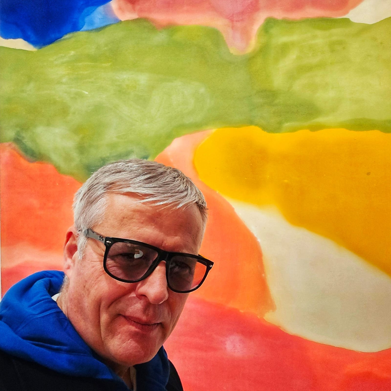 Hans Heiner Buhr, Künstler und Inhaber von Kaukasus-Reisen, posiert vor einem großformatigen, abstrakten Kunstwerk in kräftigen Rot-, Gelb- und Grüntönen.