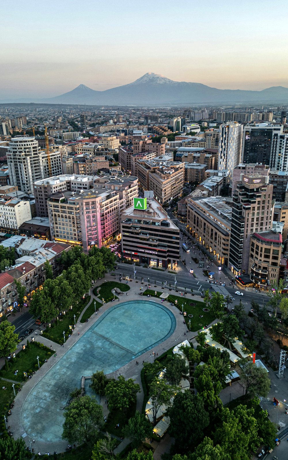 Luftaufnahme der armenischen Hauptstadt Jerewan bei Sonnenuntergang mit dem schneebedeckten Ararat im Hintergrund und dem Stadtpark im Vordergrund
