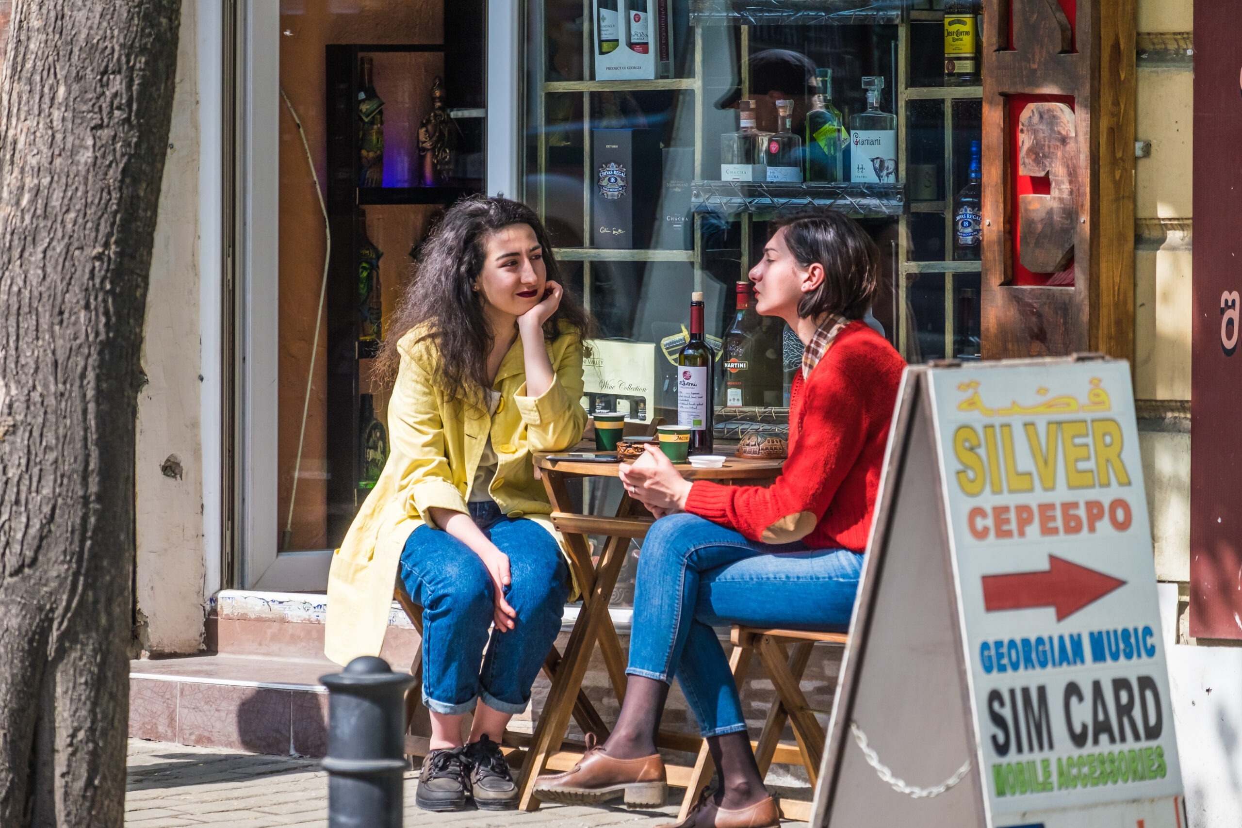 Zwei junge Frauen sitzen an einem Außentisch eines Cafés in Jerewan Armenien mit Weinflaschen und georgischer Schrift im Hintergrund