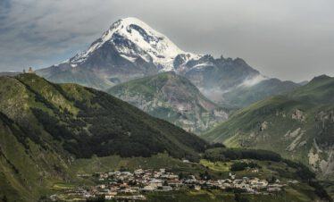 Der schneebedeckte Kasbek Gipfel mit der Dreifaltigkeitskirche Gergeti auf dem Hügel und dem Dorf Stepantsminda im Tal darunter