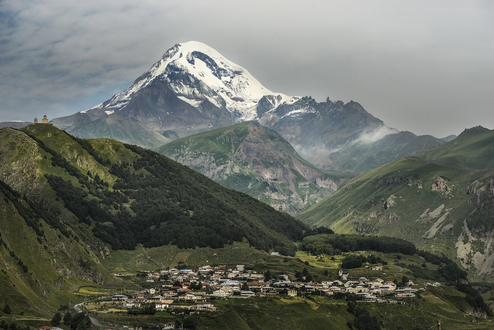 Der schneebedeckte Kasbek Gipfel mit der Dreifaltigkeitskirche Gergeti auf dem Hügel und dem Dorf Stepantsminda im Tal darunter