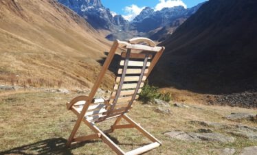 Ein leerer Holzliegestuhl auf einer herbstlichen Bergwiese mit Blick auf die Gipfel des Chaukhi-Massivs.