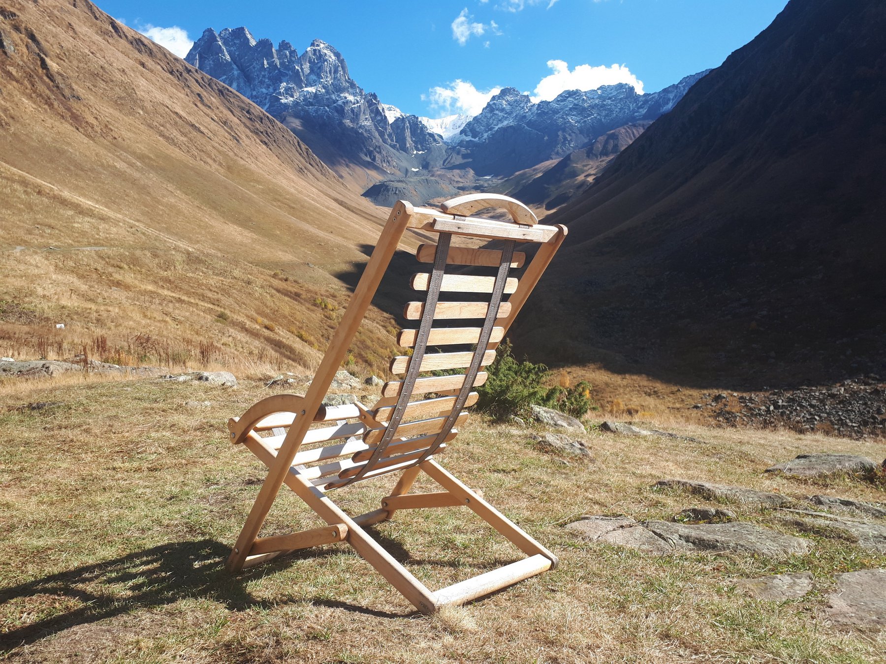 Ein leerer Holzliegestuhl auf einer herbstlichen Bergwiese mit Blick auf die Gipfel des Chaukhi-Massivs.
