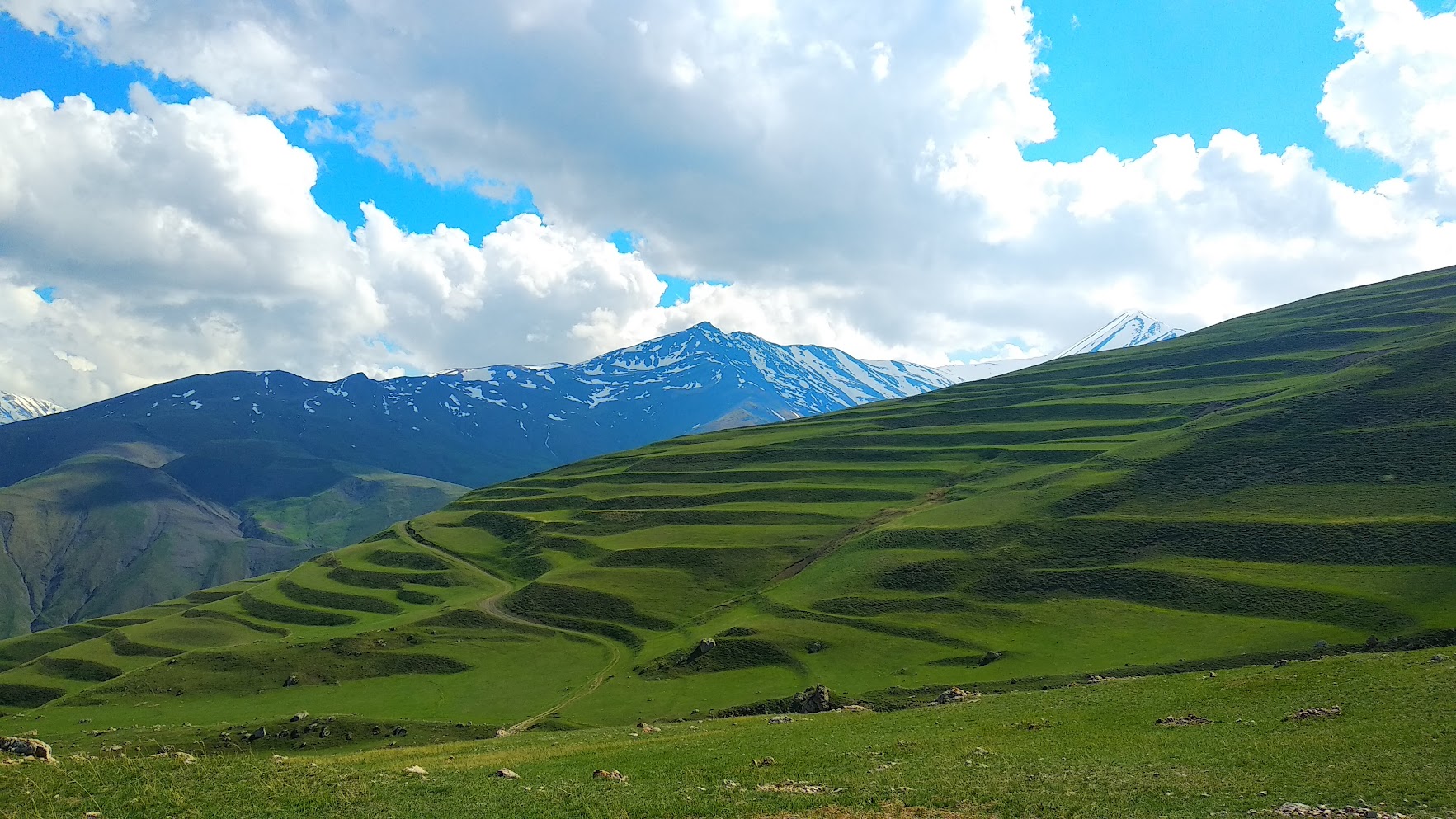 Grüne Terrassenhänge bei Khinaliq im Großen Kaukasus Aserbaidschans mit schneebedeckten Gipfeln und dramatischem Wolkenhimmel