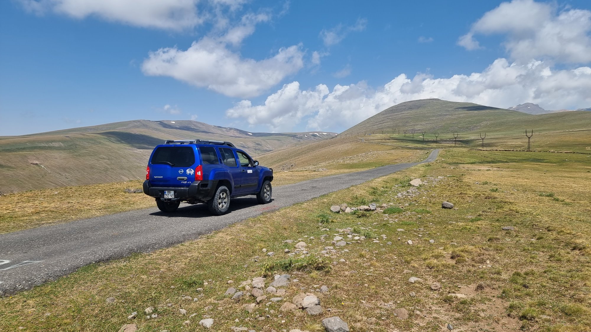 Blauer Nissan Xterra 4x4 mit Kaukasus-Reisen Logo auf einer Straße der armenischen Hochebene mit weiten Hügeln und blauem Himmel