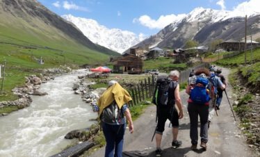 Eine kleine Reisegruppe wandert entlang eines reißenden Gebirgsflusses in Richtung eines historischen Bergdorfes in Swanetien.