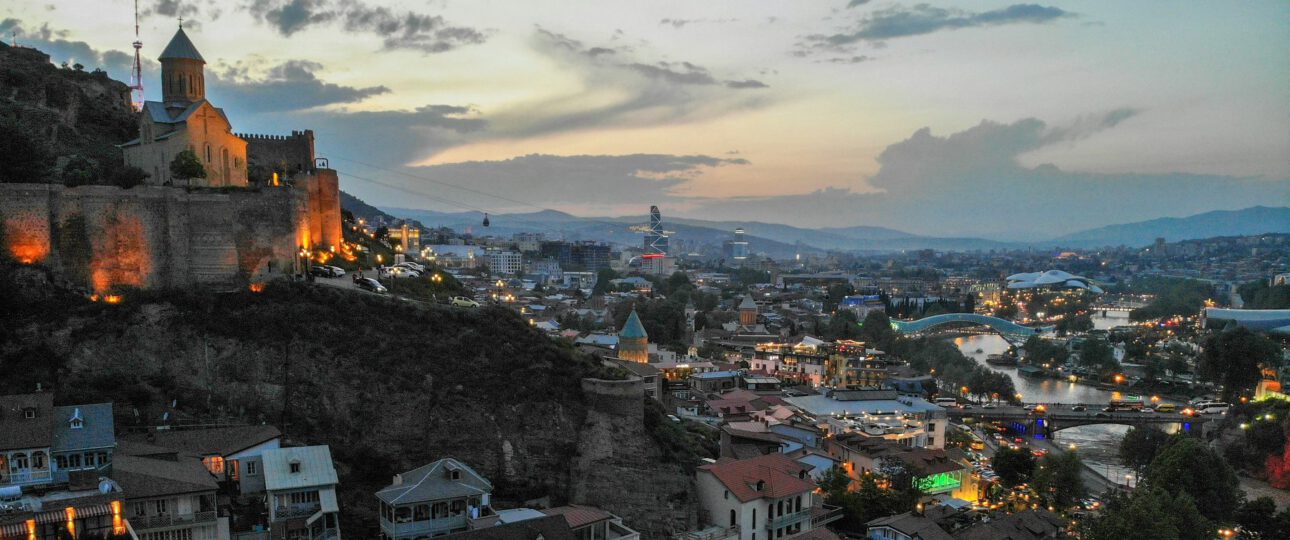 Panoramablick auf die Altstadt von Tbilissi im Abendlicht mit der beleuchteten Narikala-Festung, der Metekhi-Kirche, der Peace Bridge über den Mtkvari-Fluss und dem modernen Stadtbild im Hintergrund, Georgien