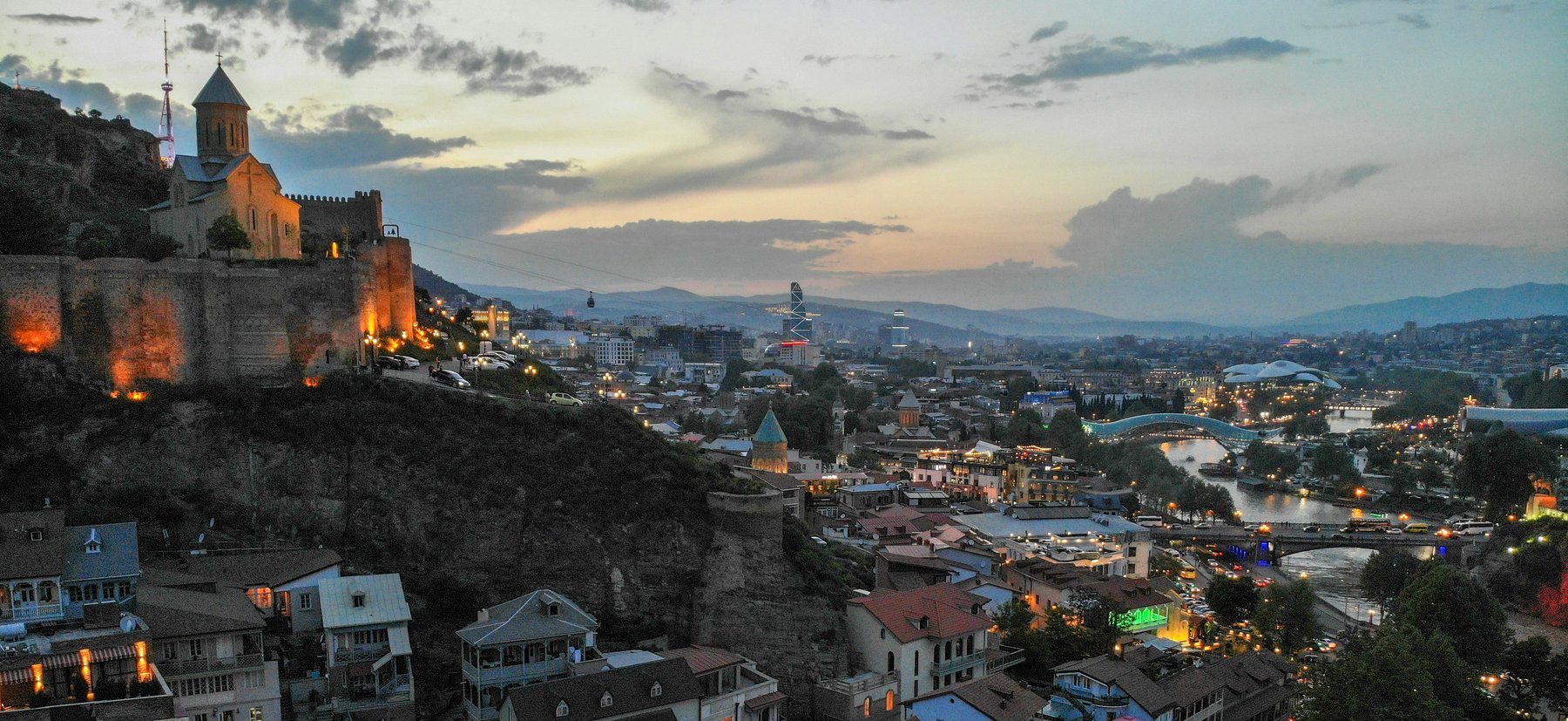 Panoramablick auf die Altstadt von Tbilissi im Abendlicht mit der beleuchteten Narikala-Festung, der Metekhi-Kirche, der Peace Bridge über den Mtkvari-Fluss und dem modernen Stadtbild im Hintergrund, Georgien