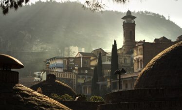 Tbilissi Altstadt im Morgendunst mit den Kuppeln der historischen Schwefelbäder und einem Minarett vor grünen Bergen