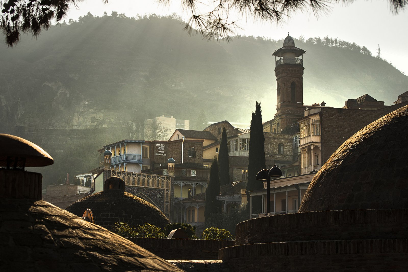 Tbilissi Altstadt im Morgendunst mit den Kuppeln der historischen Schwefelbäder und einem Minarett vor grünen Bergen