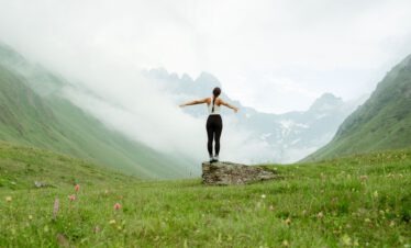 Eine Frau steht mit ausgebreiteten Armen vor der gewaltigen Bergkulisse des Kaukasus.