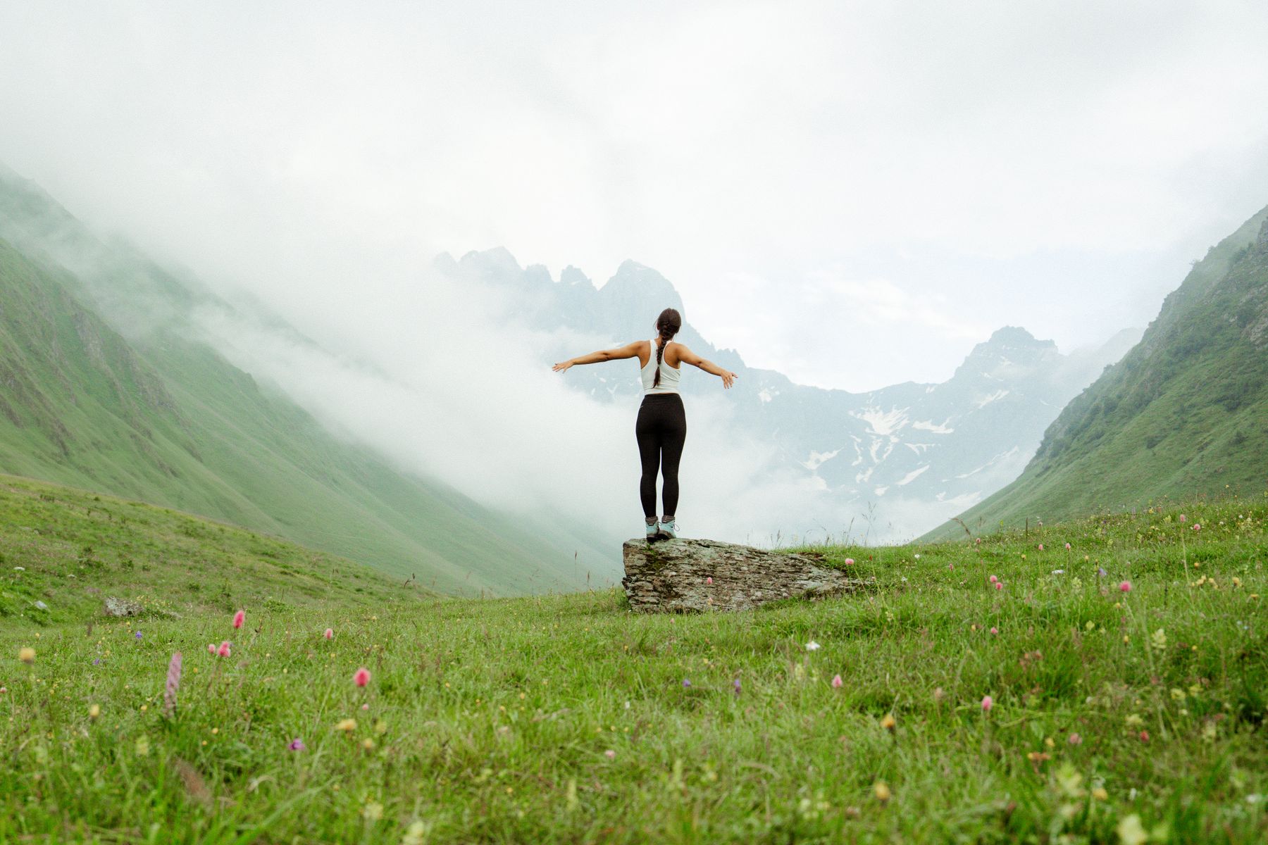 Eine Frau steht mit ausgebreiteten Armen vor der gewaltigen Bergkulisse des Kaukasus.